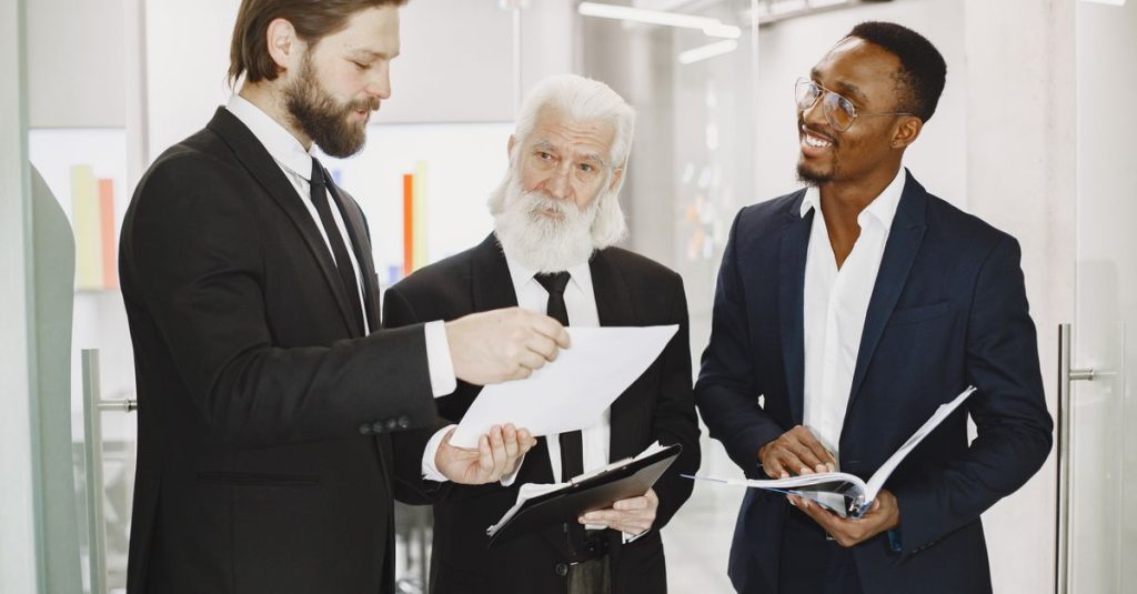 Ba người đàn ông mặc vest trao đổi tài liệu trong không gian văn phòng hiện đại.